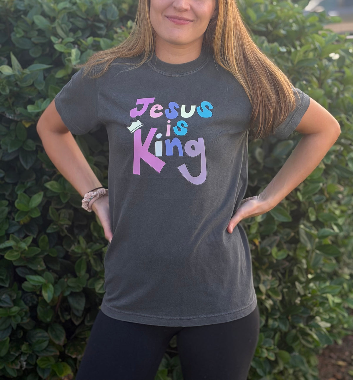 A woman wearing a dark gray graphic t-shirt with the text 'Jesus is King' in a pastel multicolor font, standing outdoors in front of a green hedge.