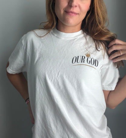A woman wearing a white crewneck t-shirt with a graphic print on the left chest that reads 'OUR GOD' in black serif font under a small gold crown, posed against a plain gray wall.