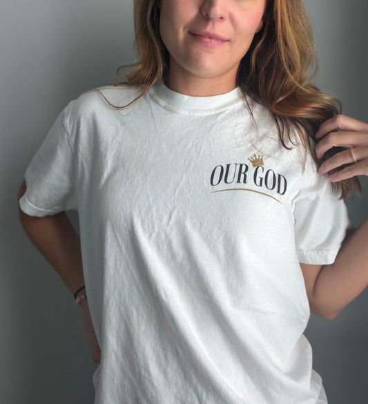 A woman wearing a white crewneck t-shirt with a graphic print on the left chest that reads 'OUR GOD' in black serif font under a small gold crown, posed against a plain gray wall.