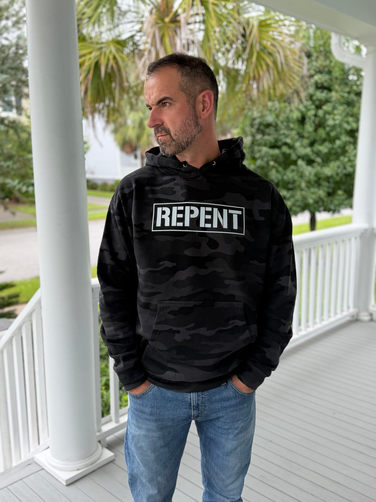 A man wearing a black camouflage hoodie with a white 'REPENT' graphic on the chest, standing on a white porch with trees in the background.
