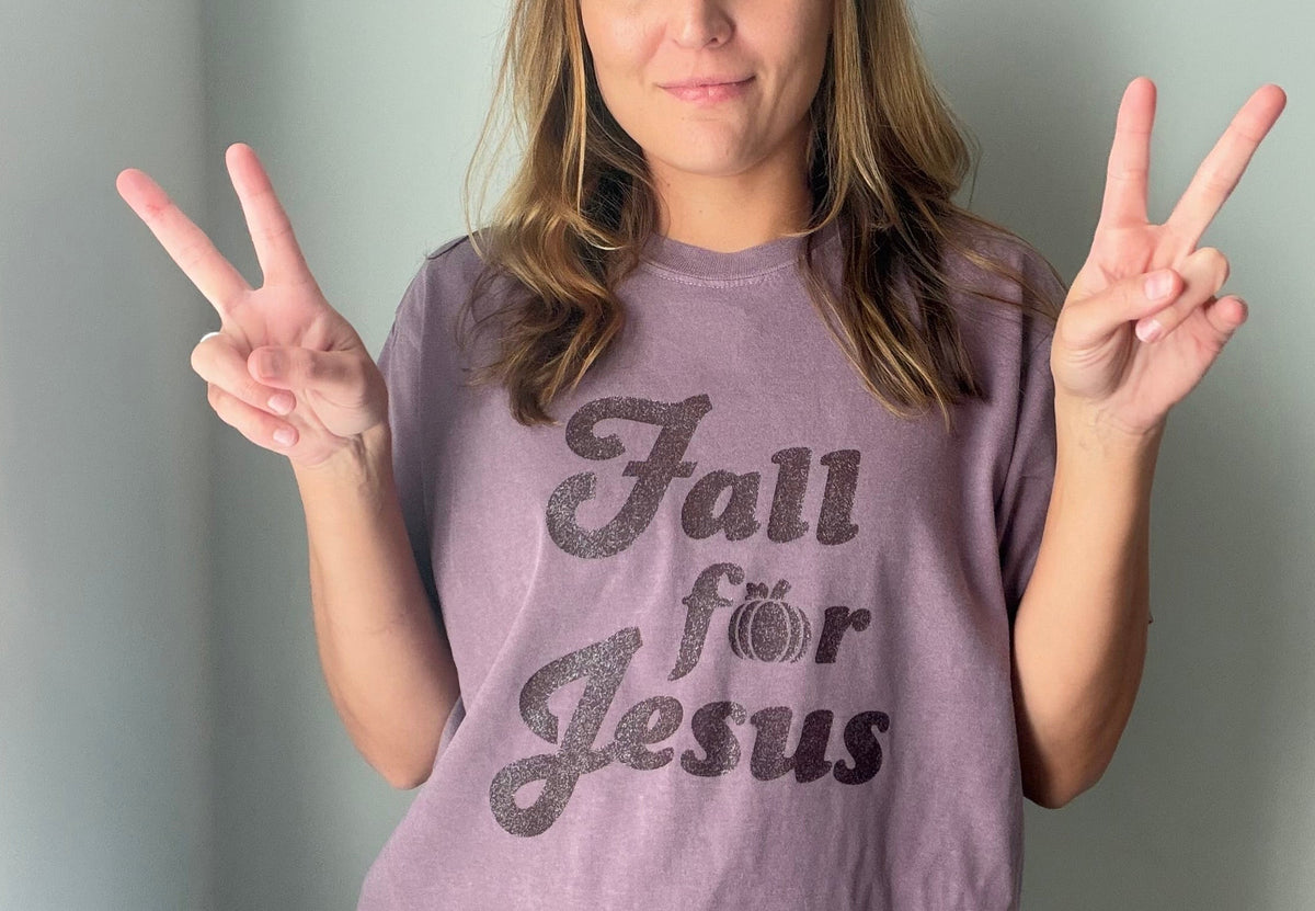 A woman wearing a lavender-purple graphic t-shirt with the text 'Fall for Jesus' in a glittery dark gray font, posing with peace signs against a plain gray wall.