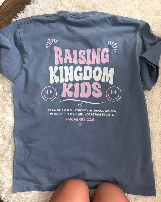 A slate blue short-sleeve t-shirt laid flat on a white shag rug, showing the back with a graphic that reads 'RAISING KINGDOM KIDS' in pink and white lettering with a Bible verse below. A person's legs are visible at the bottom edge.