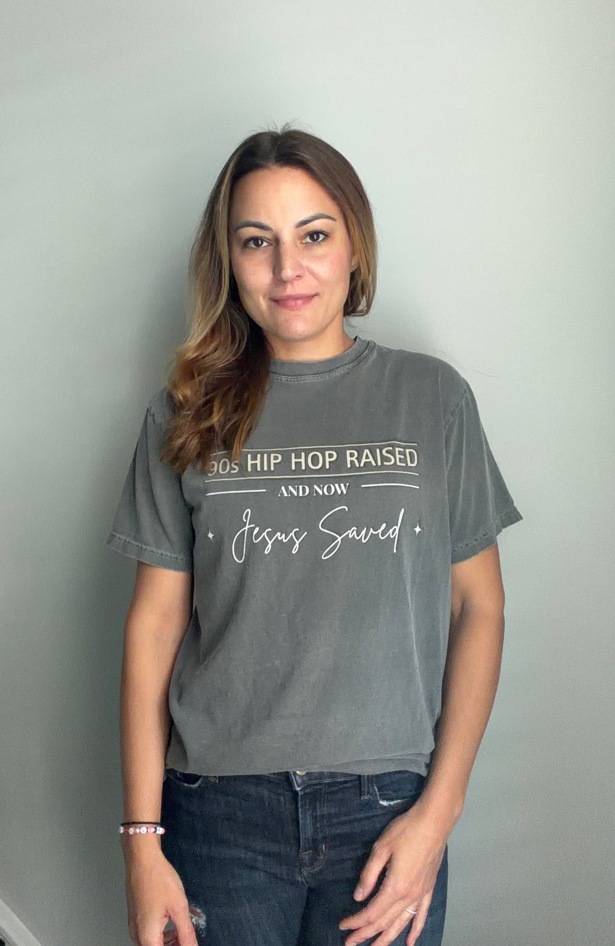A woman with long brown hair stands against a light gray wall wearing a faded dark gray graphic t-shirt with white text that reads '90s HIP HOP RAISED AND NOW Jesus Saved' and dark blue jeans.