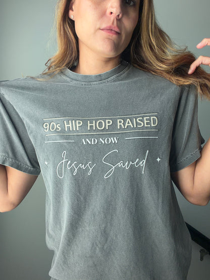 A woman wearing a faded gray graphic t-shirt with the text '90s HIP HOP RAISED AND NOW Jesus Saved' printed in white on the front, set against a plain gray wall.