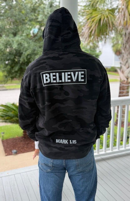 A person viewed from behind wearing a black camouflage hoodie with 'BELIEVE' printed in white inside a rectangular outline on the back, and 'MARK 1:15' near the lower back. The person is standing on a porch with trees and a railing in the background.