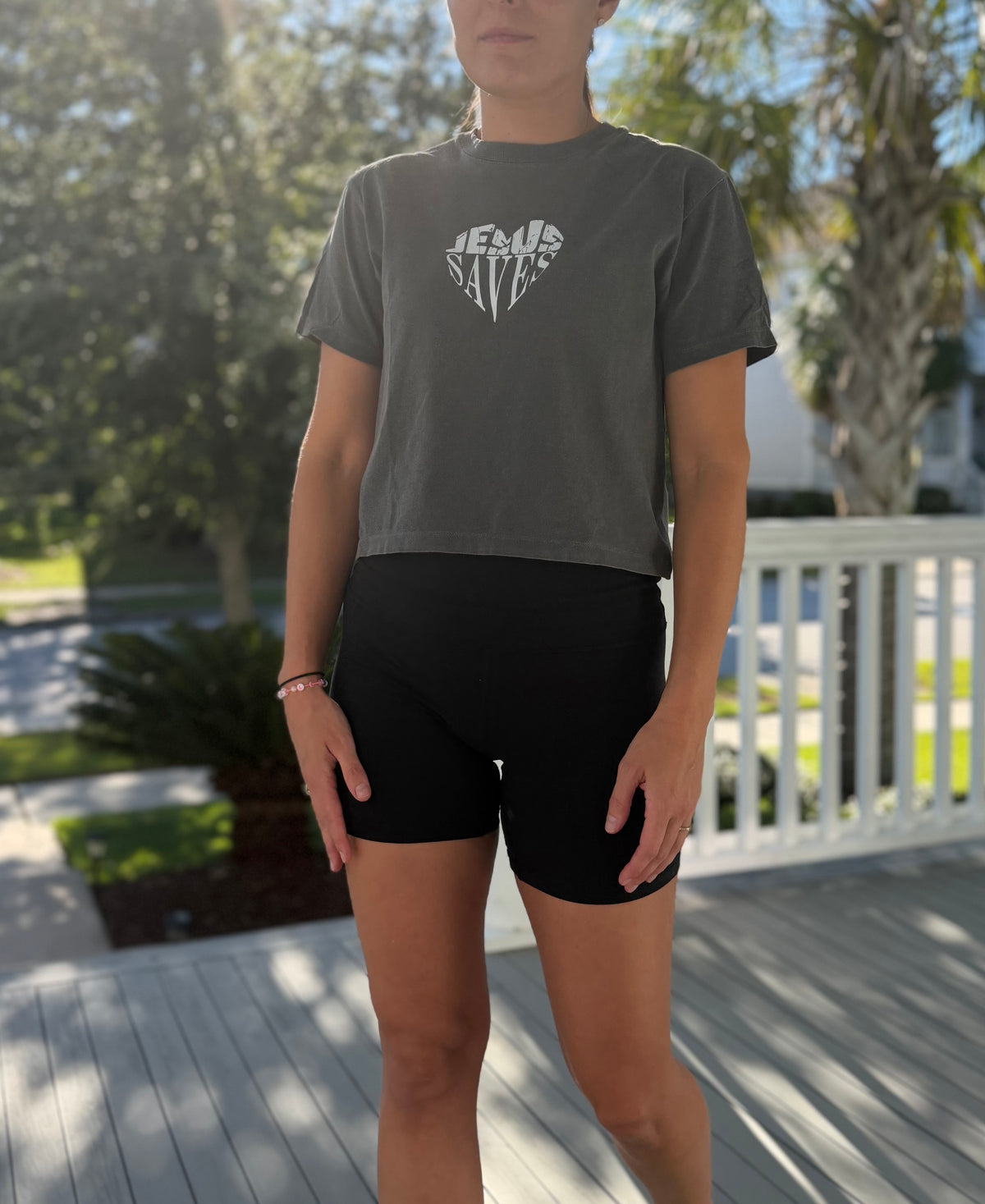 A woman wearing a dark gray graphic t-shirt with a white 'JESUS SAVES' design and black bike shorts, standing on a wooden deck outdoors.