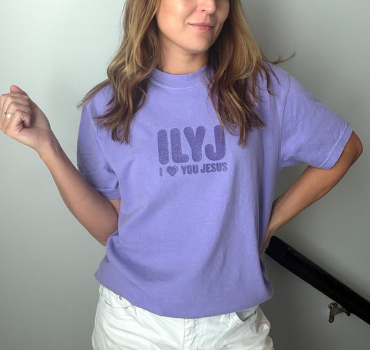 A woman wearing a lavender purple graphic t-shirt with the text 'ILYJ I ❤️ YOU JESUS' printed in a darker purple font, paired with white pants.