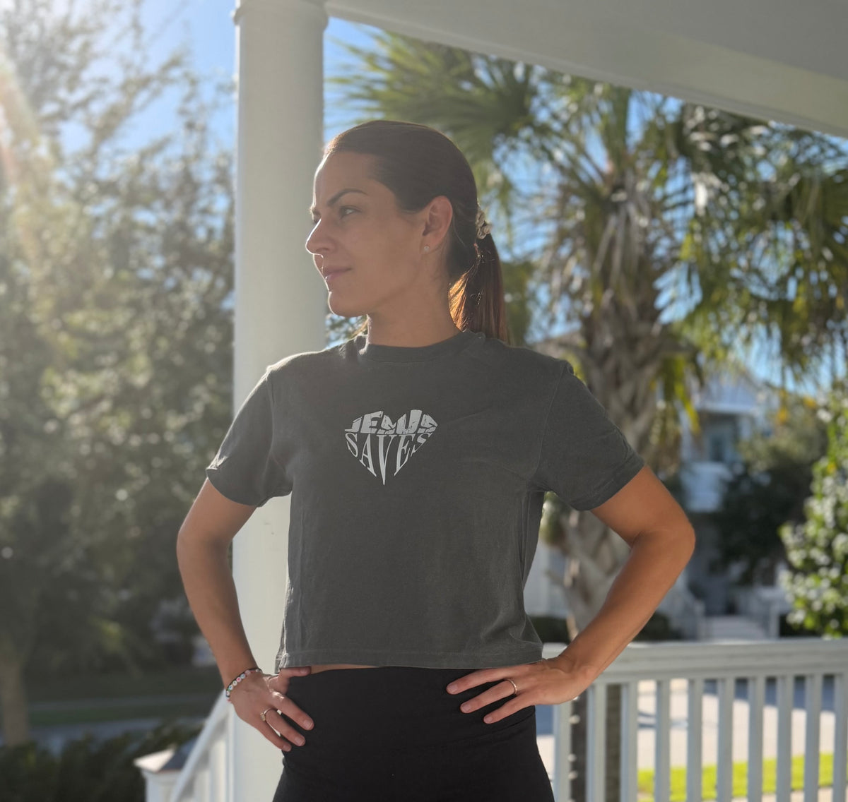 A woman wearing a dark gray graphic t-shirt with a white 'JESUS IS A ROCK' design, standing on a sunny porch with palm trees in the background.