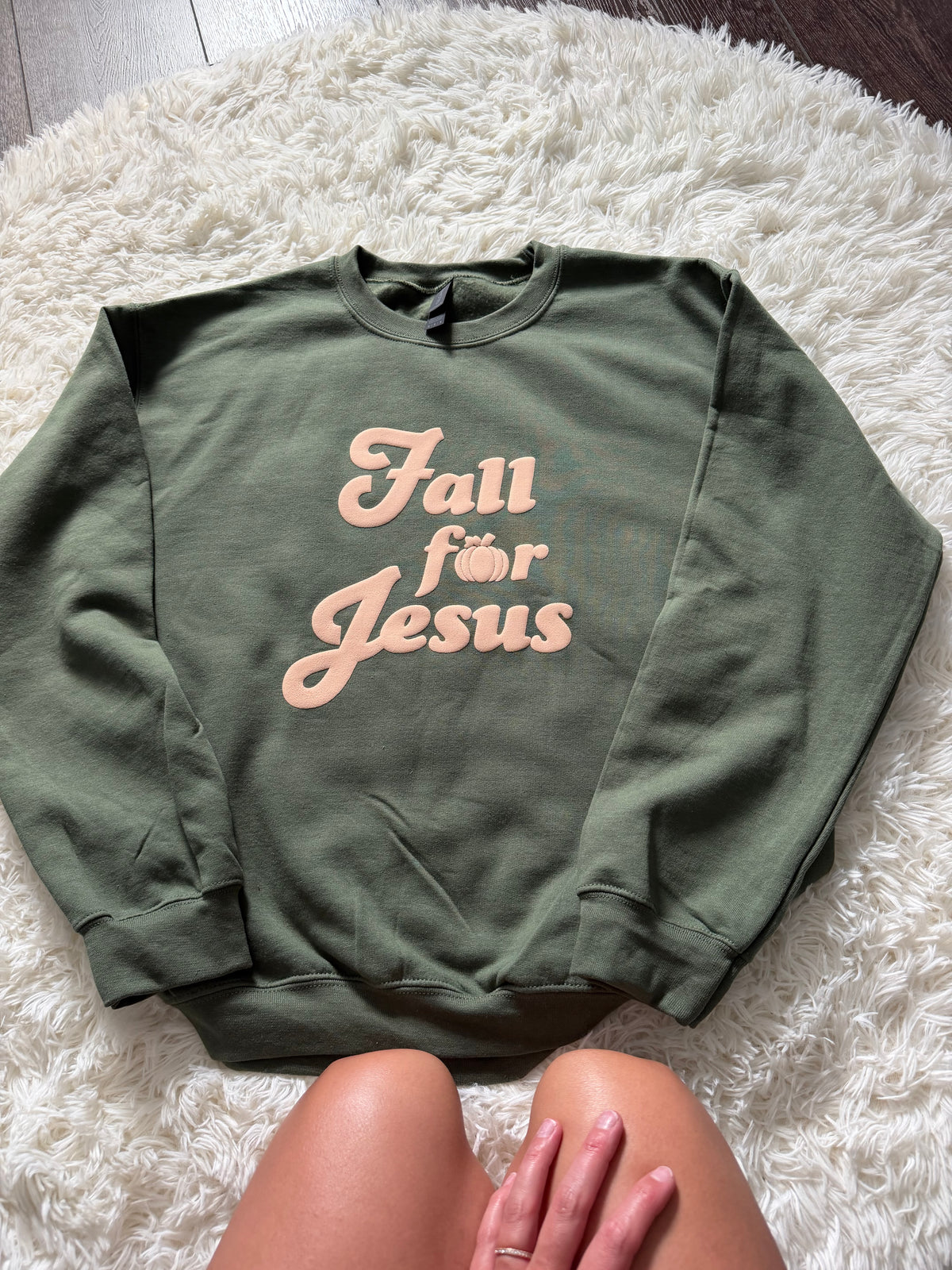 An olive green crewneck sweatshirt with the text 'Fall för Jesus' in a light peach script, laid flat on a white shag rug with a person's legs visible at the bottom.
