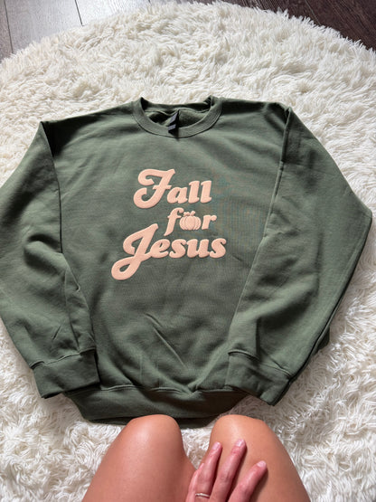 An olive green crewneck sweatshirt with the text 'Fall för Jesus' in a light peach script, laid flat on a white shag rug with a person's legs visible at the bottom.