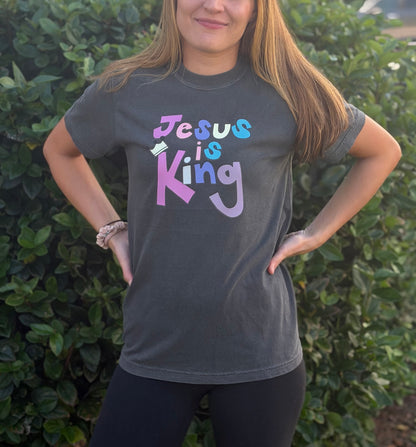 A woman wearing a dark gray graphic t-shirt with the text 'Jesus is King' in a pastel multicolor font, standing outdoors in front of a green hedge.
