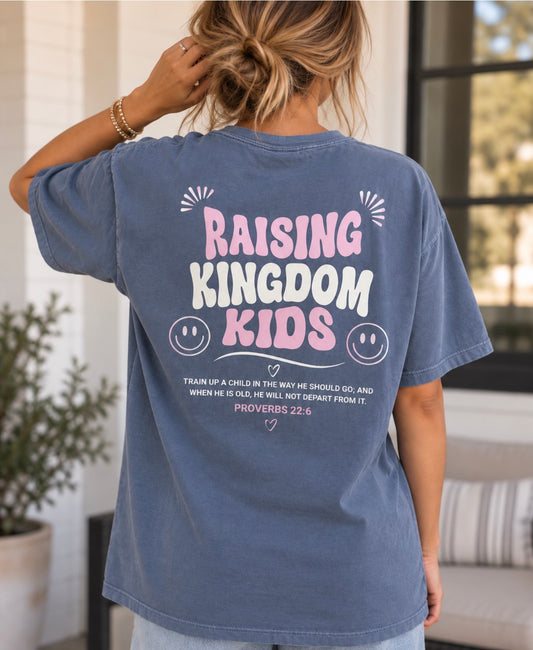A woman viewed from behind wearing a faded blue graphic T-shirt with the text 'RAISING KINGDOM KIDS' and a Bible verse printed on the back, in a home porch setting.