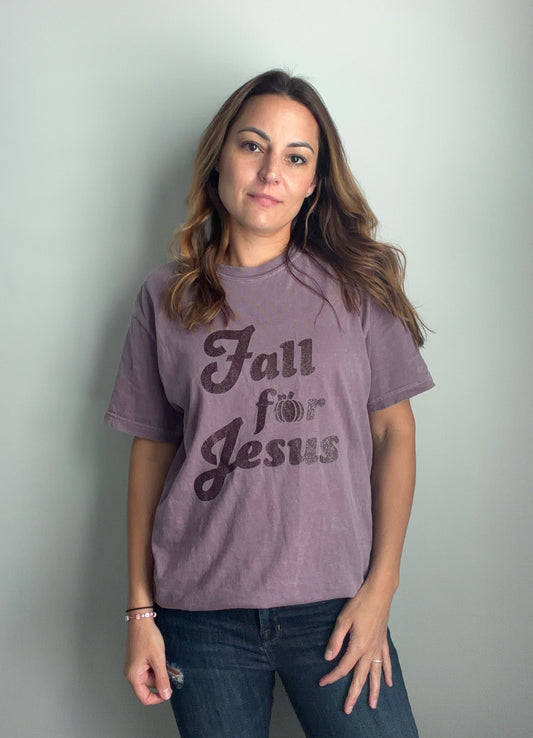A woman with long brown hair stands against a light gray wall wearing a faded mauve-purple graphic t-shirt with the text 'Fall for Jesus' in dark brown script, paired with dark blue jeans.