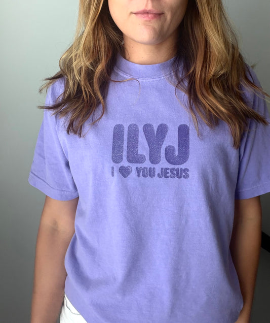 A person wearing a light lavender crewneck t-shirt with a dark purple graphic that reads 'ILYJ I ❤️ YOU JESUS' on the front, shown against a plain gray wall.