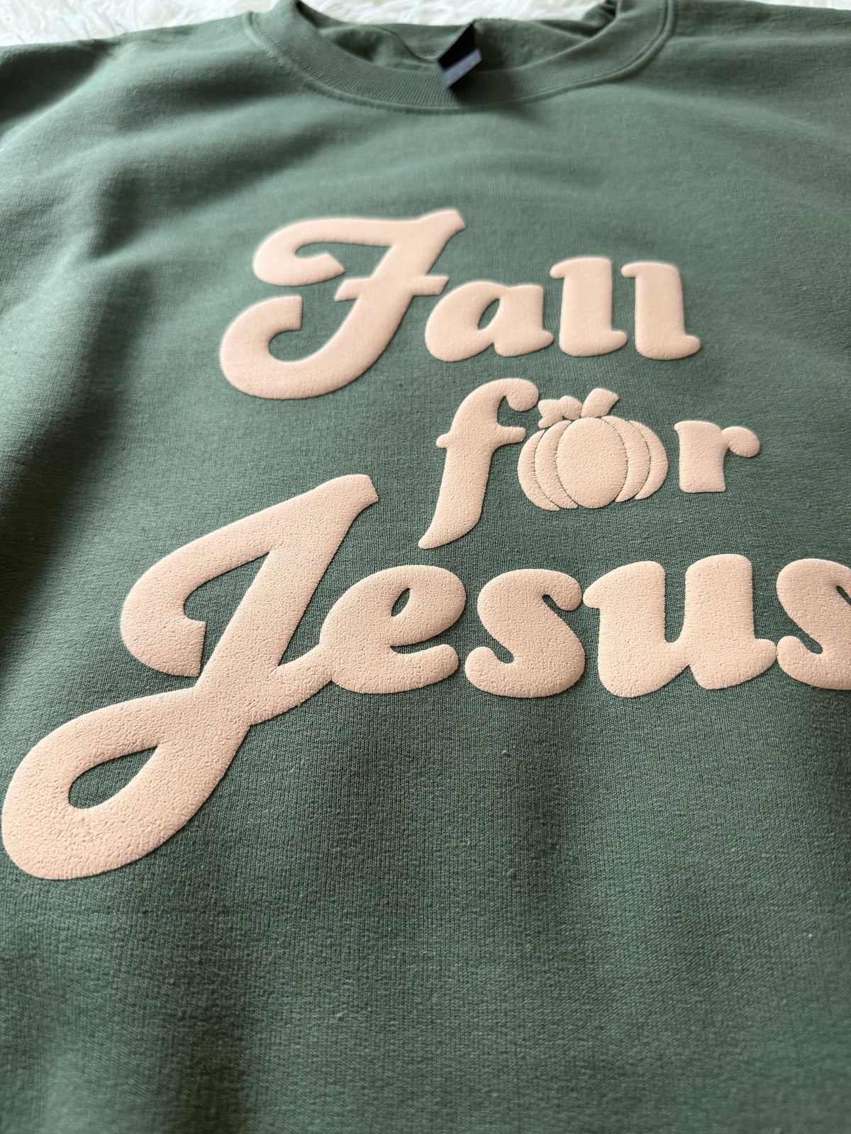 A close-up of a sage green crewneck sweatshirt featuring a raised, light beige graphic that reads 'Fall for Jesus' with a small pumpkin icon for the letter 'o'.