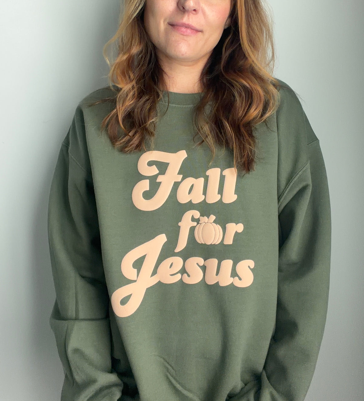 A woman wearing an olive green sweatshirt with the text 'Fall for Jesus' printed in a light peach color on the front, set against a plain light gray background.