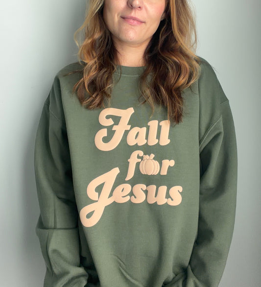 A woman wearing an olive green sweatshirt with the text 'Fall for Jesus' printed in a light peach color on the front, set against a plain light gray background.