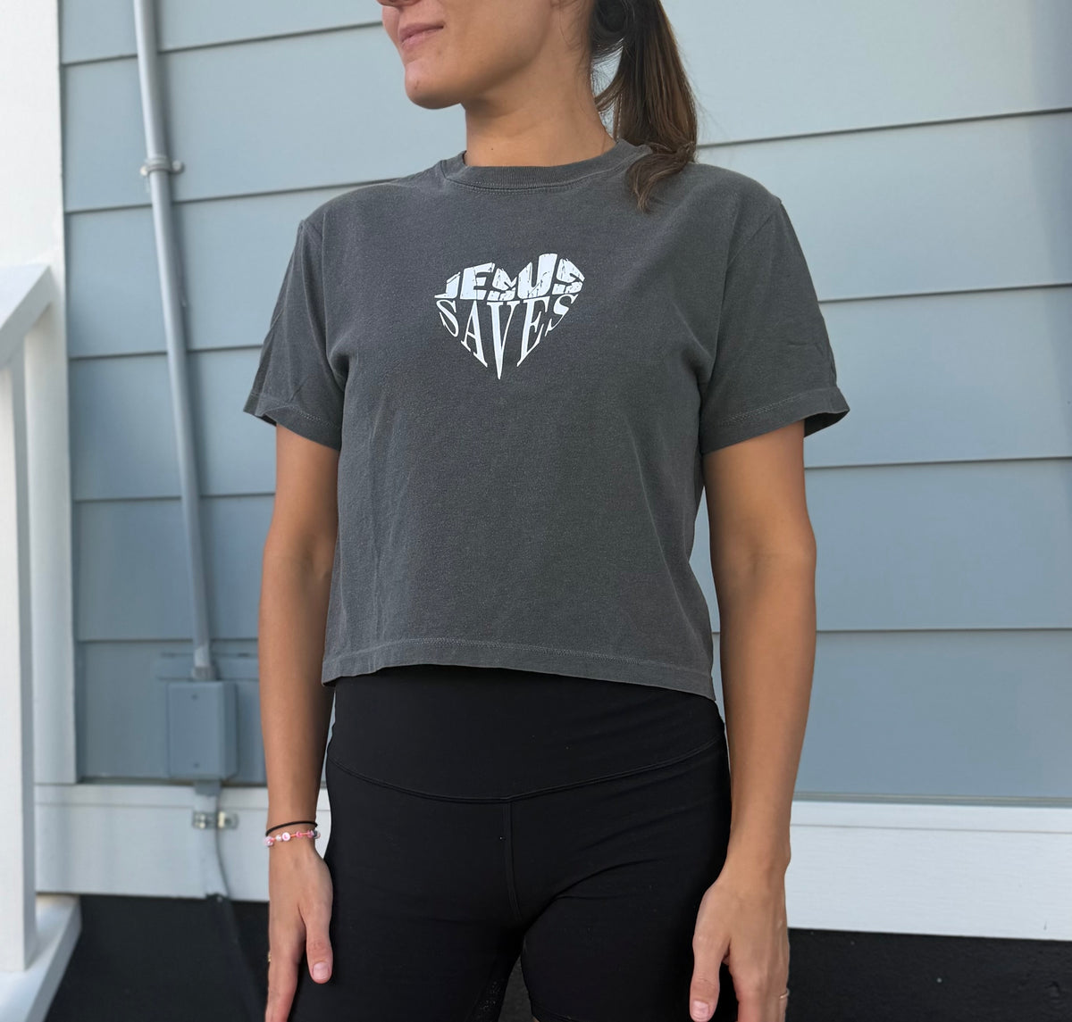 A woman wearing a dark gray cropped t-shirt with a white graphic that reads 'JESUS SAVES' in a heart shape, paired with black leggings, standing in front of a light blue-gray house siding background.