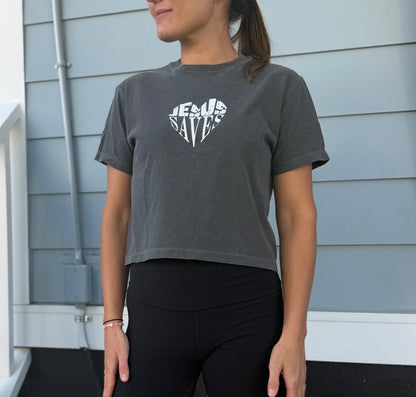 A woman wearing a dark gray cropped t-shirt with a white graphic that reads 'JESUS SAVES' in a heart shape, paired with black leggings, standing in front of a light blue-gray house siding background.