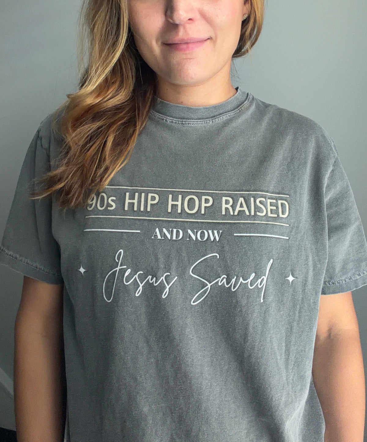 A woman with long light brown hair wearing a faded gray graphic t-shirt with white text that reads '90s HIP HOP RAISED AND NOW Jesus Saved'.