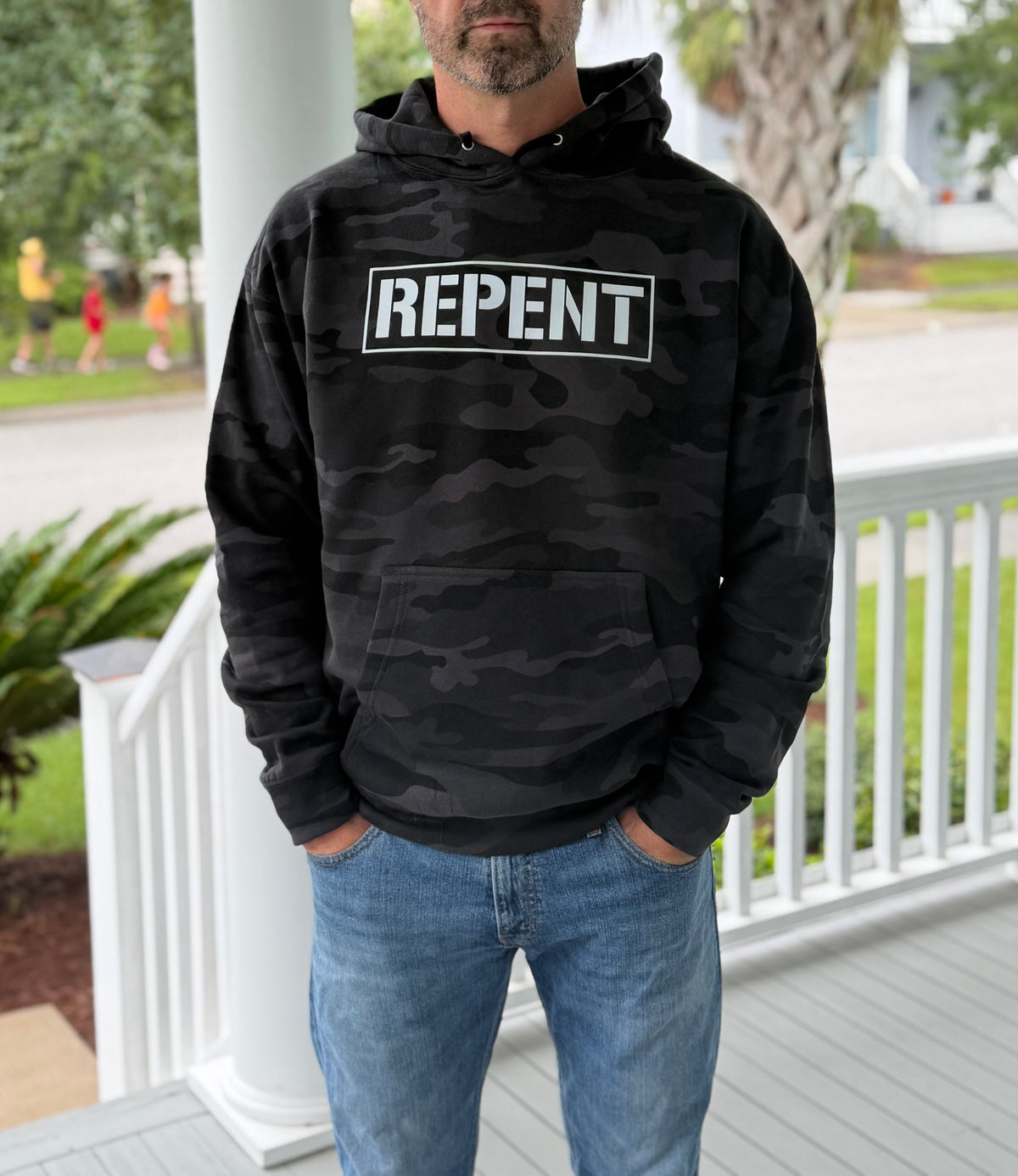 A man wearing a black camouflage hoodie with a white 'REPENT' text graphic on the chest, standing on a white porch.