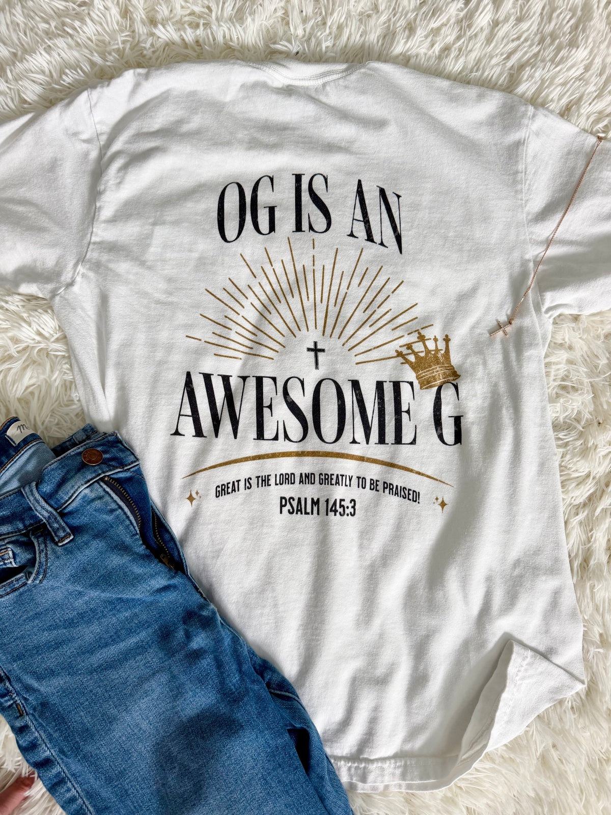 A white graphic T-shirt laid flat on a white faux-fur surface, styled with blue jeans. The shirt features a black and gold design with the text 'OG IS AN AWESOME G', a cross, sunburst, small crown, and 'PSALM 145:3'.