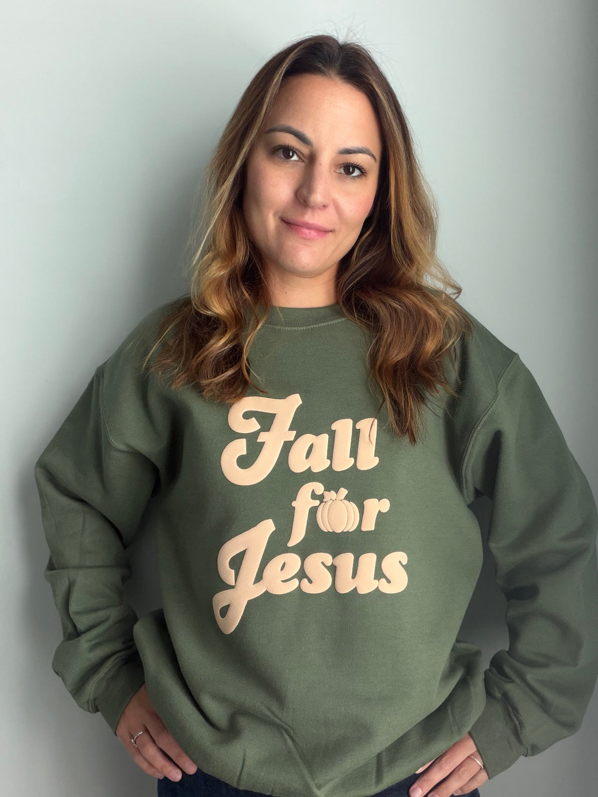 A woman with shoulder-length brown hair wearing an olive green crewneck sweatshirt with the text 'Fall for Jesus' printed in a beige script font on the front, posing against a light gray wall.