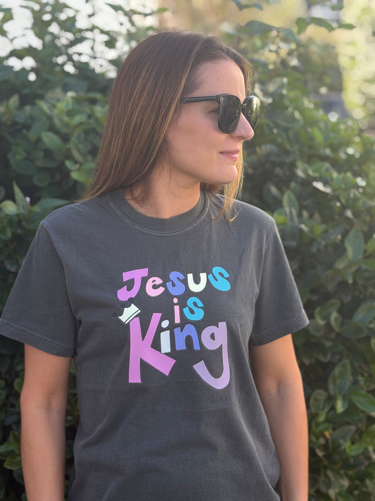 A woman wearing a dark gray graphic t-shirt with the text 'Jesus is King' in a colorful pastel font, standing outdoors in front of green foliage.