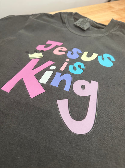 A dark gray heather t-shirt featuring a colorful graphic print that reads 'Jesus is King' in pink, blue, yellow, and purple lettering, with a small gold crown icon above the 'J'.