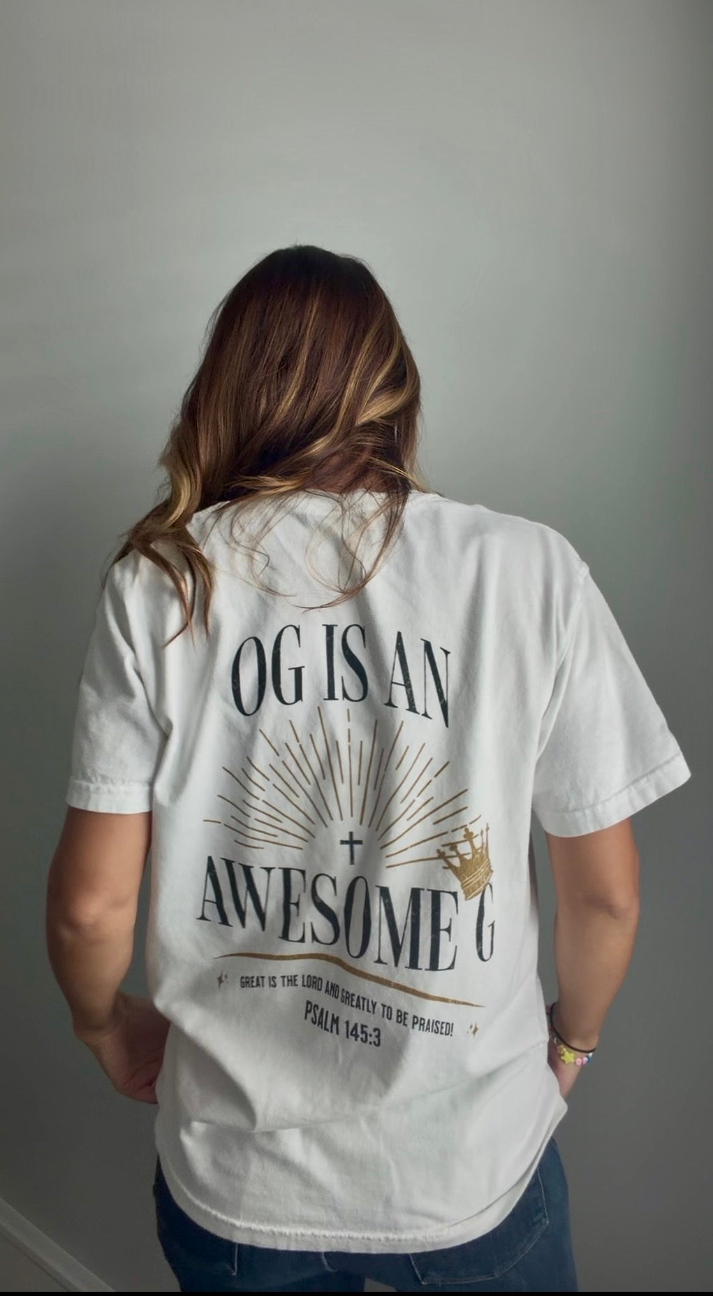 A woman viewed from behind wearing a white graphic t-shirt with 'OG IS AN AWESOME G' printed in black and gold on the back, paired with blue jeans against a plain gray wall.
