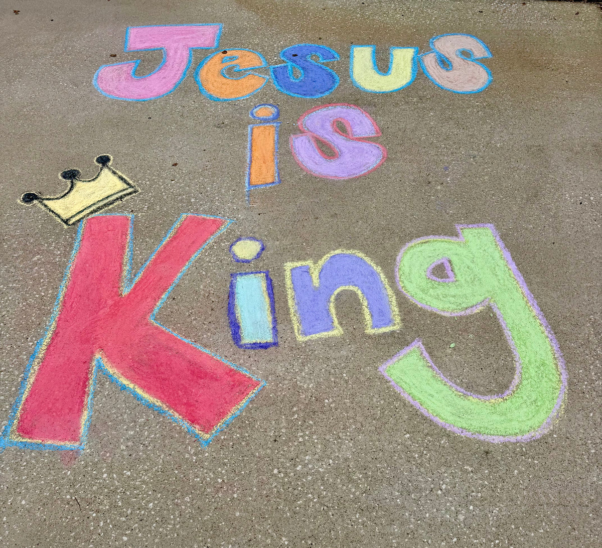 Colorful chalk lettering on concrete pavement that reads 'Jesus is King,' with the word 'King' in large pink, blue, and green letters and a small gold crown drawn near the letter 'J'.